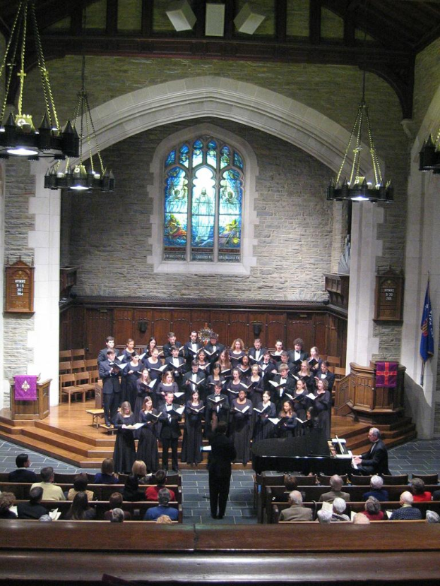 Eine Gruppe von Menschen, die in der Mitte stehen und Bücher halten, mit Stühlen dahinter, andere sitzen unten, Kronleuchter an der Decke, eine Flagge rechts und ein Fenster oben.