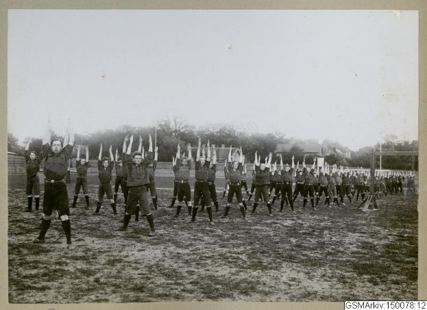 Schwarzes und weißes Foto einer Fußballmannschaft in Militäruniformen aus den 1930er Jahren, die ihre Hände auf einem Feld mit Bäumen und einem klaren Himmel in der Luft hält.