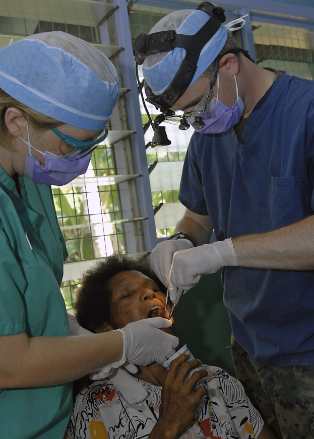Zwei Chirurgen in Masken und Handschuhen führen eine Gesichtsoperation an einer Frau durch, die auf einem Bett liegt, mit Bäumen im Hintergrund durch ein Fenster sichtbar