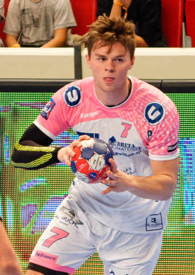 Ein Mann in einem pinken Hemd mit einem Fußball, daneben die Hand einer anderen Person und einige Menschen im Hintergrund auf Stühlen sitzend.