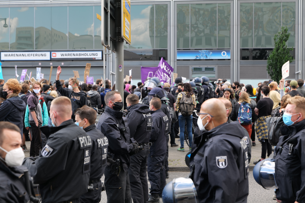 Eine große Gruppe von Menschen steht vor einem Gebäude, einige halten Schilder und tragen Helme, mit einem Mast mit einer Schildertafel im Vordergrund und einem Baum im Hintergrund, scheinbar protestierend.