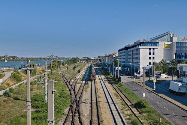 Ein Zug fährt auf Schienen neben einer Stadtlandschaft mit Gebäuden, Fahrzeugen, Bäumen und Infrastruktur unter einem bewölkten Himmel.
