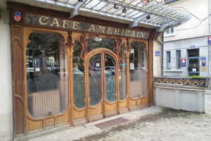 Ein Café in Paris mit Glasfenstern und -türen, einem Schild mit Text, Deckenleuchten und einer Leitung an der Gebäudeseite.