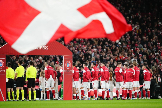 Gruppe von Menschen auf einem Fußballfeld mit einer roten und weißen Fahne im Vordergrund, einem Bogen mit der Aufschrift "Bayern München vs Bayern München Wett-Tipps & Vorschau" im Hintergrund und einer großen Menge im Stadion.