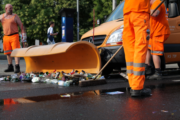 Eine Gruppe von Männern in orangefarbenen Uniformen reinigt Müll am Straßenrand, mit einem Schneepflug im Vordergrund und verschiedenen Fahrzeugen, Polen, Bäumen und einer Person mit einem Besen im Hintergrund, zusammen mit einem nahen Müllcontainer.