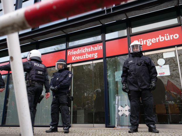 Eine Gruppe von Polizisten in Helmen und Handschuhen steht vor einem Glaswand-Gebäude, mit einem Pfahl links und Schildern im Hintergrund, die "Friedrichshain Kreuzberg" und Papiere an den Glaswänden angeklebt zeigen.