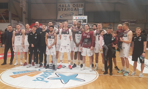 Gruppe von Männern und Kindern auf einem Basketballfeld mit Schildern "Hala Osir Starga", Zuschauern, einem Basketballkorb, Metallstangen, Deckenlampen und einer Wand im Hintergrund.