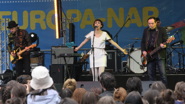 Eine Bühne mit vier Musikern: eine Frau in einem weißen Kleid singt in der Mitte, zwei Männer spielen Gitarren auf der linken und rechten Seite und eine Menge vorne mit einer Wand und Lautsprechern im Hintergrund.