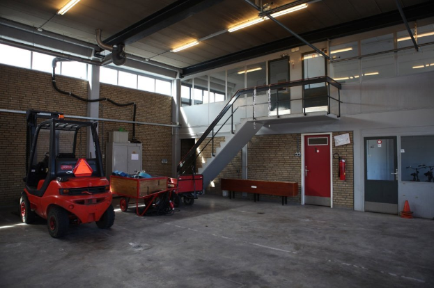 Ein großes Lagerinnere mit einem roten Gabelstapler, einem Wagen mit verschiedenen Gegenständen, einer Bank, einem Feuerlöscher, einer Treppe mit Geländern, Türen, Fenstern, Rohren, Deckenleuchten und einem Rad.