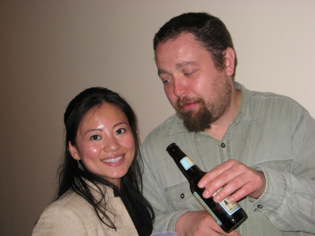 Ein Mann mit einer Flasche starrt eine lächelnde Frau an, beide stehen vor einer Wand.