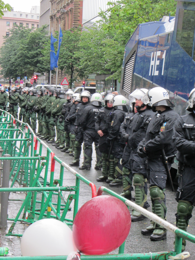 Eine Gruppe von Polizisten in Helmen und mit Waffen steht vor einer Barriere auf einer Straße mit Geländern und Luftballons, mit Bäumen, Gebäuden, Flaggen und einem klaren blauen Himmel im Hintergrund.