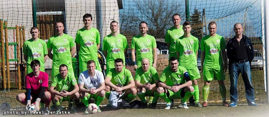 Gruppe von Männern in grünen T-Shirts auf einem Fußballfeld mit einem Ball und einem Torpfosten, Hintergrund enthält Bäume, Gebäude und Fahrzeuge.