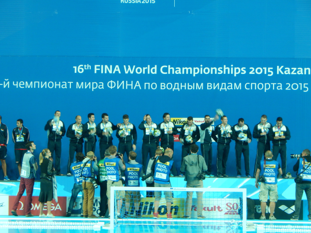 Eine Gruppe von Menschen steht oben auf einem Swimmingpool, einige halten Kameras, mit einem Netz im Vordergrund und einem Banner mit der Aufschrift "FINA World Championships 2015 Kazan" im Hintergrund.