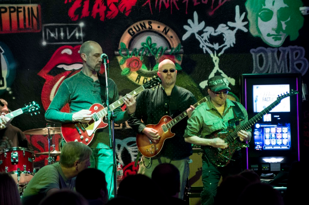 Drei MÃ¤nner, die auf einer BÃ¼hne Gitarren spielen, mit einer Menge darunter und einem Graffiti oder einer Wandmalerei im Hintergrund.