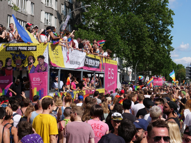 Eine große Menschenmenge marschiert während des Berlin Pride 2019 die Straße entlang, viele tragen Kappen und Schutzbrillen, einige halten Fahnen, mit Bannern, Laternenmasten, Bäumen und Gebäuden im Hintergrund bei bewölktem Himmel.