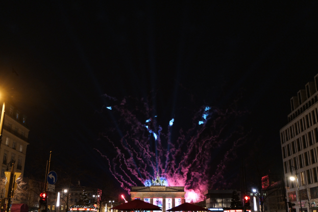 Eine belebte Stadtstraße in Berlin, Deutschland, mit beleuchteten Gebäuden, Pfählen, Schildern, Verkehrszeichen, Zelten und Fahrzeugen, mit dem Brandenburger Tor im Hintergrund unter einem Feuerwerk-erhellten Nachthimmel.