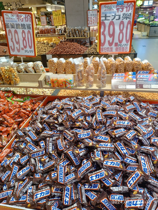 Ein Ladenvitrine mit verschiedenen Süßigkeiten, einschließlich Snickers-Riegeln, neben anderen Lebensmitteln und Hintergrund-Elementen.