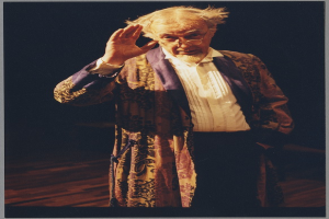 Ein Mann in einer Robe und Brille steht auf einer Bühne mit dunklem Hintergrund und schaut ernsthaft ins Publikum.