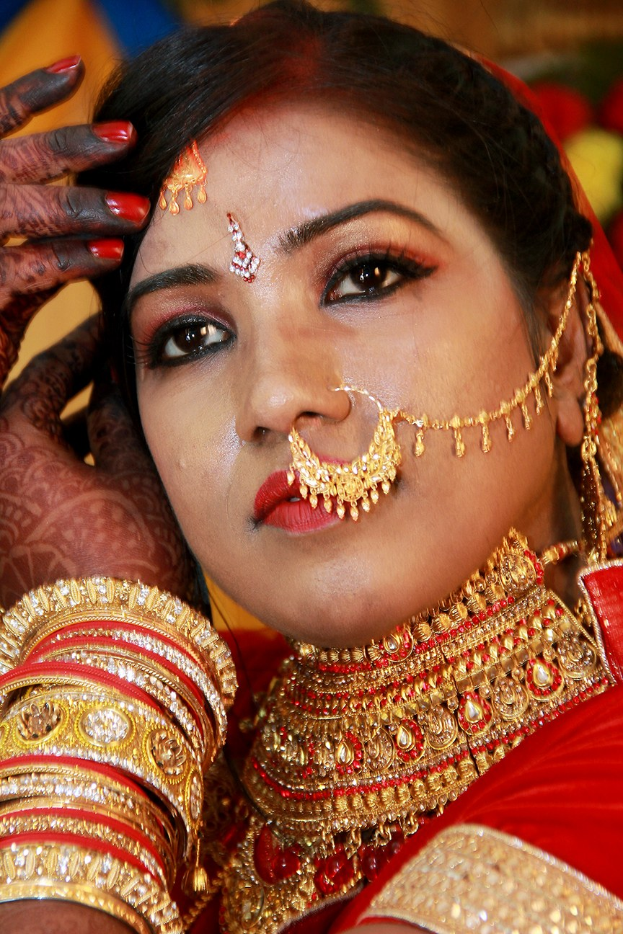 Eine Frau in einem roten und goldenen Brautoutfit mit traditionellen Schmuckstücken und Armreifen, die vor einem unscharfen Hintergrund steht.