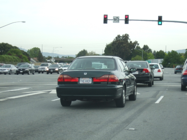 Eine befahrene Straße mit mehreren Fahrzeugen, drei Strommasten und einer Ampel auf der rechten Seite bei klarem Himmel.