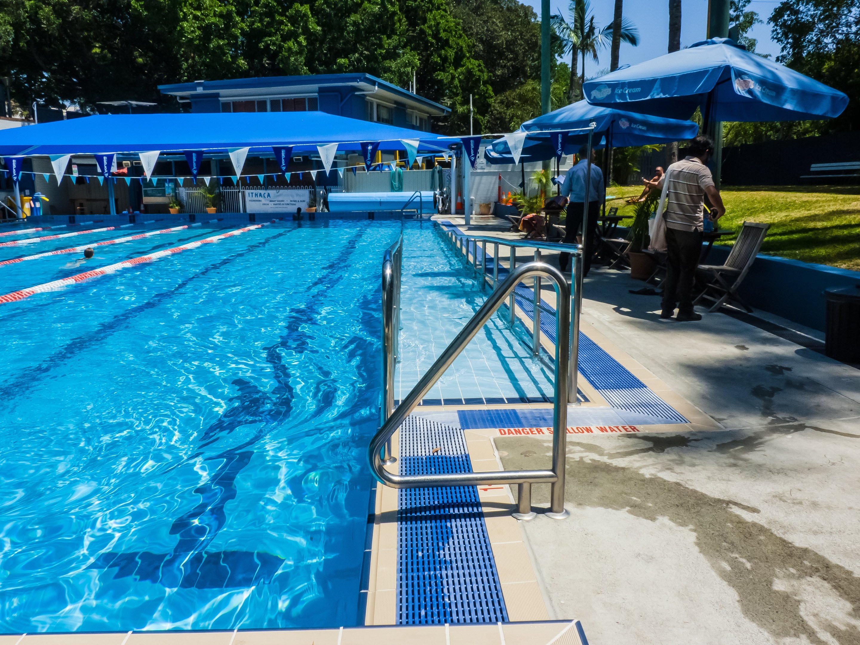 Großes Freibad mit schwimmenden Menschen, umgeben von Geländern, Sonnenschirmen, Stühlen, Tischen, Pflanzen, Gras, Bäumen und einem Gebäude unter einem klaren blauen Himmel.