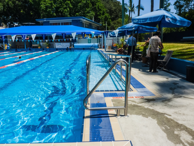 Großes Freibad mit schwimmenden Menschen, umgeben von Geländern, Sonnenschirmen, Stühlen, Tischen, Pflanzen, Gras, Bäumen und einem Gebäude unter einem klaren blauen Himmel.
