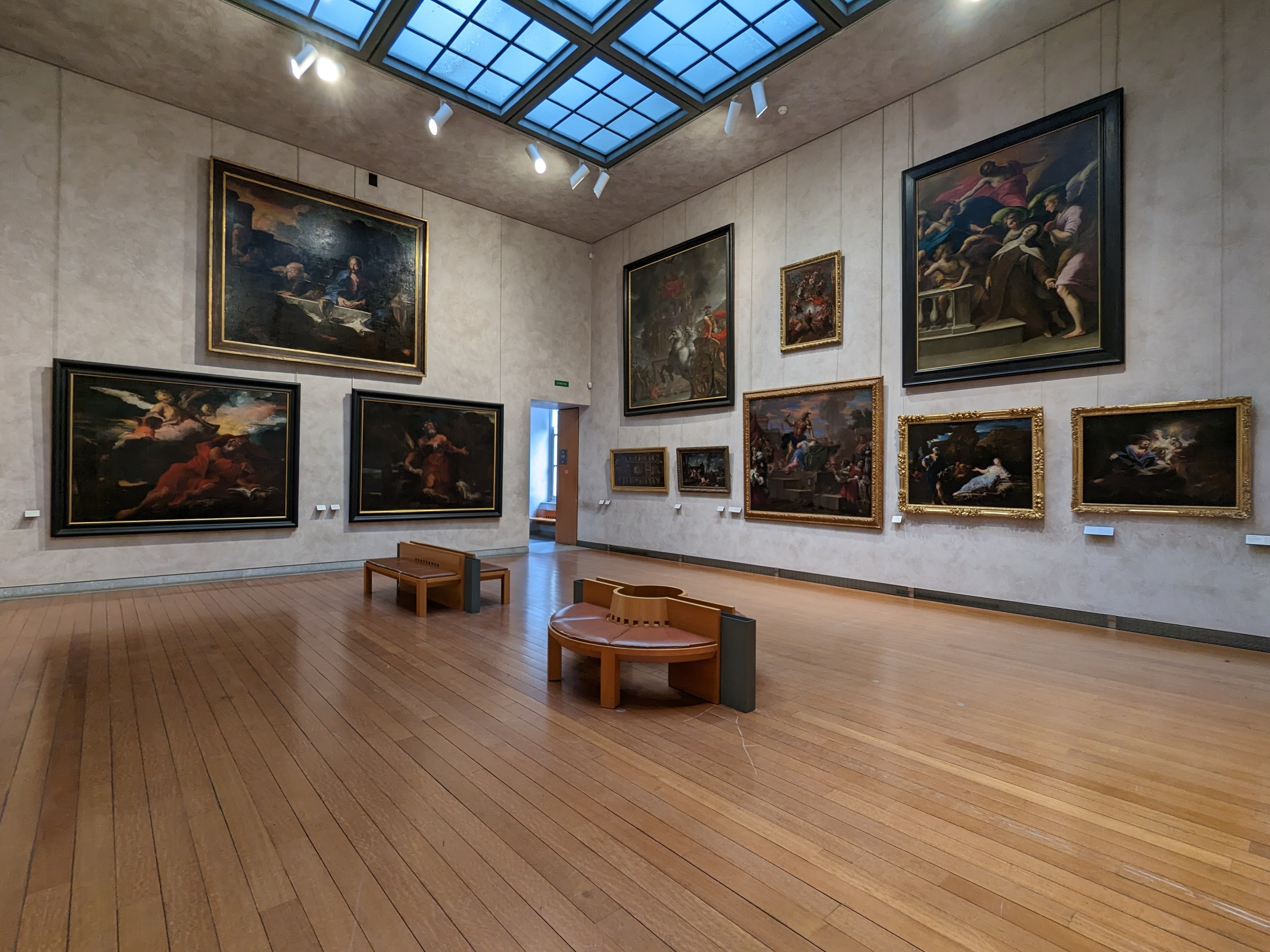 Eine geräumige Kunstgalerie mit zahlreichen Gemälden an den Wänden, hölzernen Bänken auf dem Boden und Deckenleuchten, gelegen in der National Gallery of Art in London.
