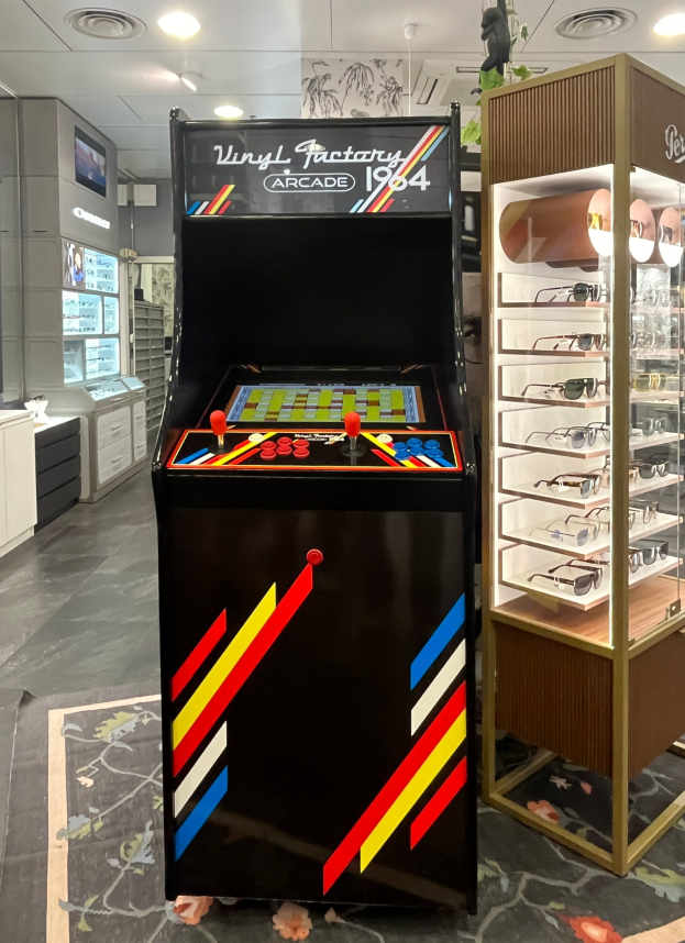 Ein Geschäft mit einem zentralen Retro-Arcade-Automaten, einer Brillenauslage auf der rechten Seite und einer retro-stylischen Beleuchtung mit Schränken, Fernseher und anderen Gegenständen im Hintergrund.