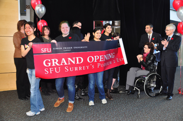 Eine Gruppe von Frauen hält ein Banner, daneben stehen zwei klatschende Männer und eine Frau im Rollstuhl, die aufgeregt aussieht, vor einem schwarzen Vorhang mit Ballons im Hintergrund.