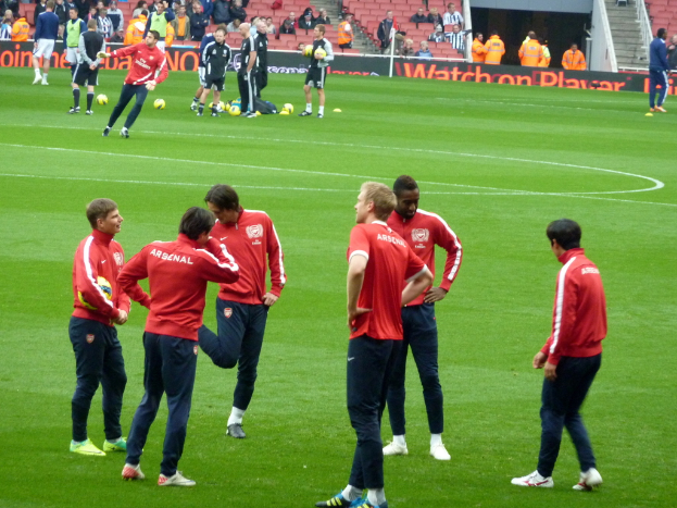 Eine Gruppe von Fußballspielern auf einem grünen Feld mit Fußballen und einer Zuschauerschaft im Hintergrund auf Stühlen sitzend.