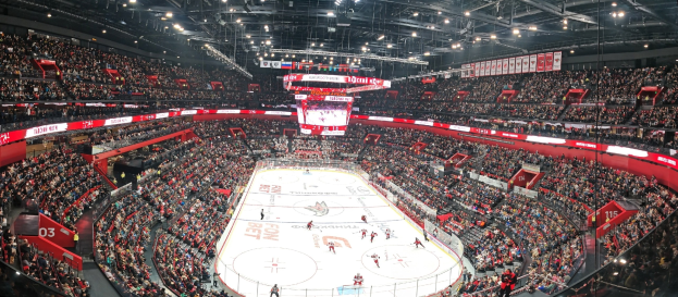 Eine große Indoor-Hockey-Arena mit Zuschauern auf den Rängen, heller Deckenbeleuchtung und einem zentralen Scoreboard-Bildschirm.