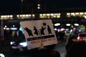 Eine Person hält ein Schild mit der Aufschrift "Berlin gegen Nazis" vor einer Menge und Fahrzeugen, mit einem beleuchteten Gebäude im Hintergrund.