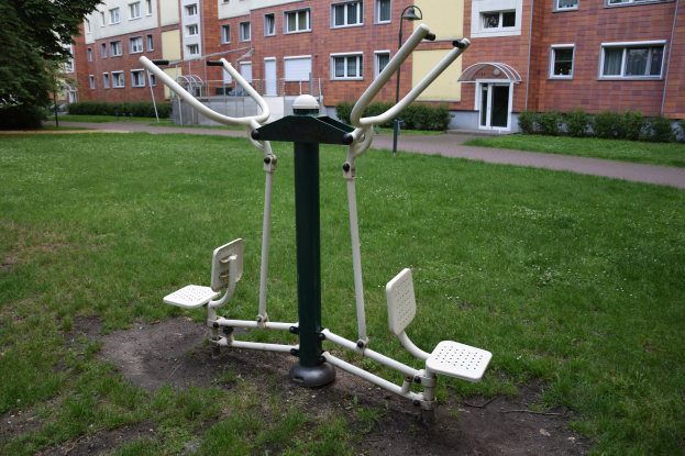 Outdoor-Gym-Ausrüstung auf einer grünen Wiese mit Bäumen, Pflanzen und Gebäuden im Hintergrund.