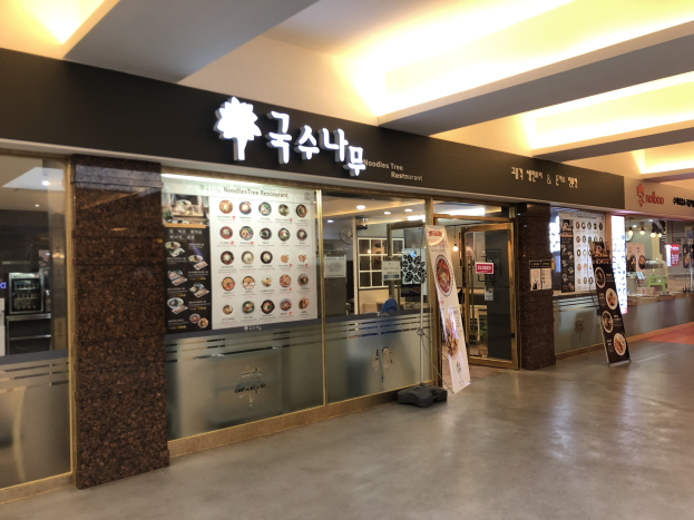 Der Eingang zum Noodles Tree Restaurant in einem Einkaufszentrum, mit Glaswänden und -türen, beleuchteter Decke und Menschen auf der rechten Seite.