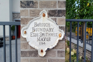 Ein Steinplakette an einem Gebäude trägt die Inschrift "Gemeindegrenze Edm Smithwick Bürgermeister", mit einem Geländer auf der rechten Seite, Fahrzeugen auf der linken Seite und Gebäuden, Pflanzen und Bäumen im Hintergrund.