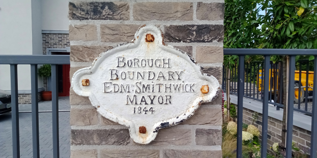 Ein Steinplakette an einem Gebäude trägt die Inschrift "Gemeindegrenze Edm Smithwick Bürgermeister", mit einem Geländer auf der rechten Seite, Fahrzeugen auf der linken Seite und Gebäuden, Pflanzen und Bäumen im Hintergrund.