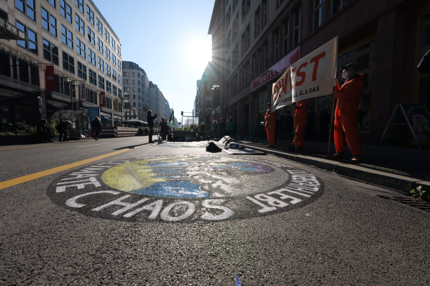 Menschen mit Schildern und Plakaten stehen auf einer Straße, die von Pflanzen und Gebäuden gesäumt ist, mit "Chaos" auf die Straße gemalt in Bezug auf die Occupy Wall Street Demonstration.