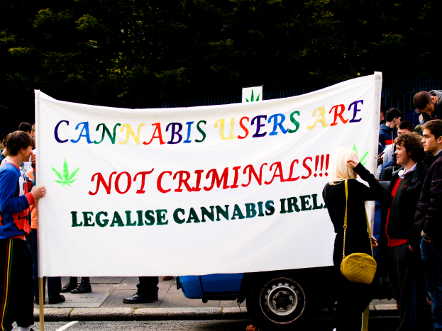Eine Gruppe von Menschen steht auf der Straße und hält ein Banner mit der Aufschrift "Cannabisnutzer sind keine Kriminellen", daneben ein parkendes Fahrzeug und im Hintergrund Bäume und ein Zaun.
