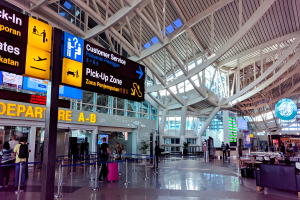 Ein Flughafen-Terminal-Inneneinrichtung mit Menschen, die gehen, einige tragen Taschen, und Pfosten mit Bändern; Sofas und Tische mit Gegenständen auf der rechten Seite und ein Glaswand-Gebäude mit Deckenleuchten im Hintergrund.