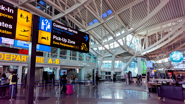 Ein Flughafen-Terminal-Inneneinrichtung mit Menschen, die gehen, einige tragen Taschen, und Pfosten mit Bändern; Sofas und Tische mit Gegenständen auf der rechten Seite und ein Glaswand-Gebäude mit Deckenleuchten im Hintergrund.