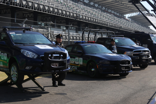 Eine Gruppe von Polizeiwagen vor einer Rennstrecke geparkt, uniformierte Personen auf der Straße, Stadion und Maschendrahtzaun im Hintergrund.