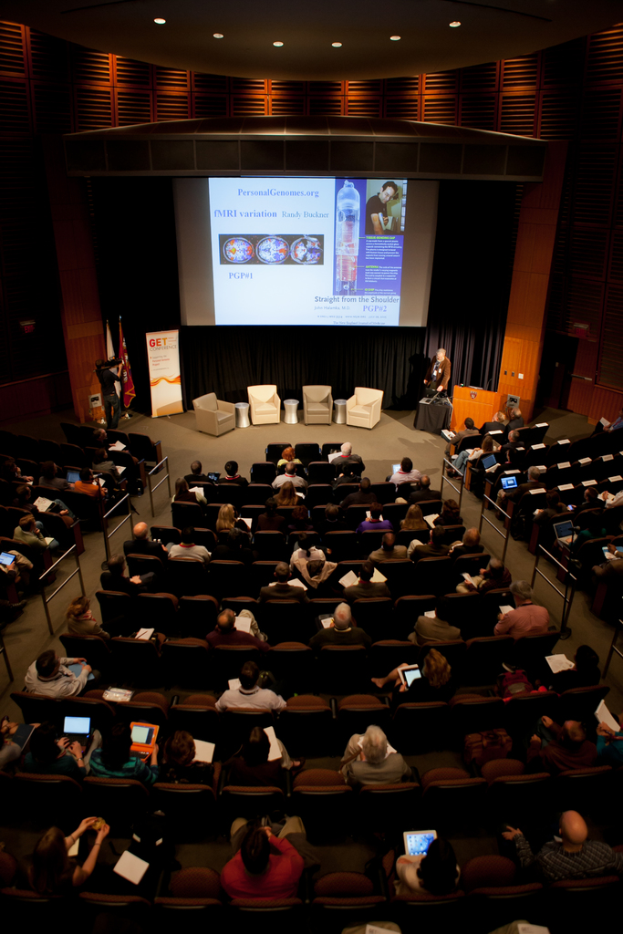 Ein mit Sitzenden Menschen, die Gegenstände halten, beleuchteter Auditoriumssaal mit Flaggen links, einem Podium rechts und einem Werbeplakat links.