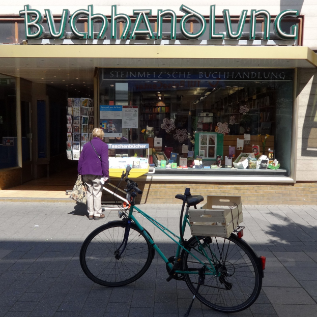 Eine Frau steht neben einem Fahrrad, das vor einem Laden geparkt ist, mit einem Fahrradrad im Vordergrund und durch die Glastür des Ladens sichtbaren Büchern.