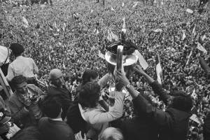 Schwarzes und weißes Foto einer Gruppe von Menschen, die einen Pokal in die Höhe halten, vor einer großen Menge, einige mit Kameras und Fahnen.