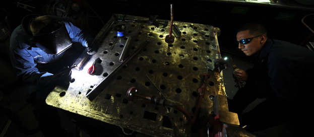 Zwei Männer in Schutzausrüstung arbeiten an einem Metallstück in einer dämmrig beleuchteten Werkstatt, umgeben von verschiedenen Werkzeugen und Materialien auf einem Tisch.