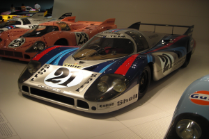 Ein Porsche 917K Le Mans Rennwagen wird in einem Museum ausgestellt, umgeben von anderen Sportwagen und Menschen, mit einer Wand im Hintergrund und einem Blatt Papier mit Text links daneben.