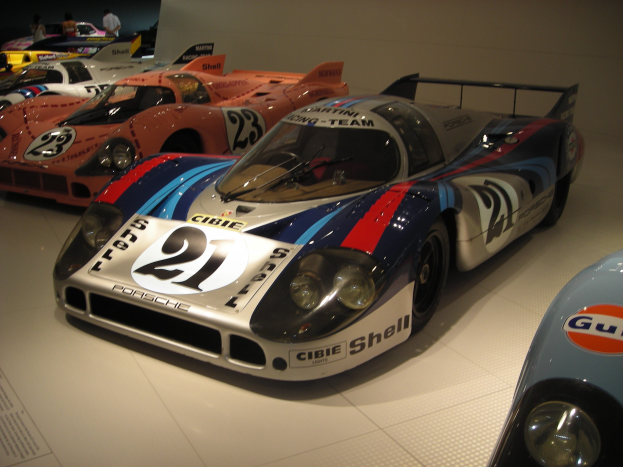 Ein Porsche 917K Le Mans Rennwagen wird in einem Museum ausgestellt, umgeben von anderen Sportwagen und Menschen, mit einer Wand im Hintergrund und einem Blatt Papier mit Text links daneben.