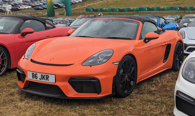 Ein Porsche 718 Boxster GTS auf dem Goodwood Festival of Speed, zusammen mit anderen Autos, einem Banner, Bäumen und einem klaren blauen Himmel im Hintergrund.