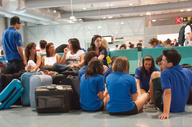 Ein belebtes Flughafeninnere mit wartenden Passagieren, Gepäck zu links und Glastüren mit Richtungsschildern im Hintergrund.