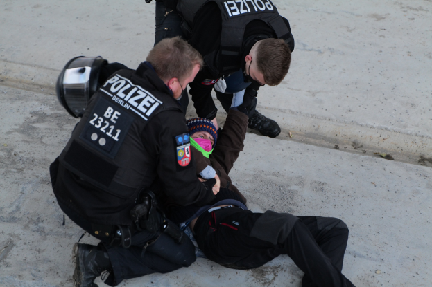 Ein Mann liegt auf dem Boden mit zwei Polizeibeamten daneben, einer hält ein Kind; der Mann trägt eine Mütze und die Beamten haben Text auf ihren Jacken.